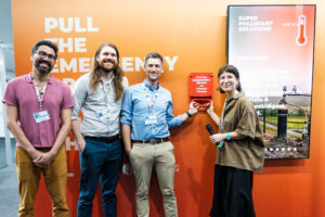 Four panelists stand smiling in front of an orange display at a climate conference. Two of them are holding a red device labeled ‘Pull the emergency brake on climate change,’ symbolically activating it. A screen beside them shows information about methane and pollutant solutions. The group appears to have just finished a panel on methane and public health.