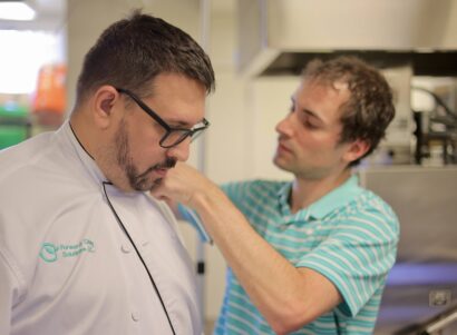 A scientist setting up a clear tube on a chef's shoulder to test working exposures in a kitchen while he cooks.